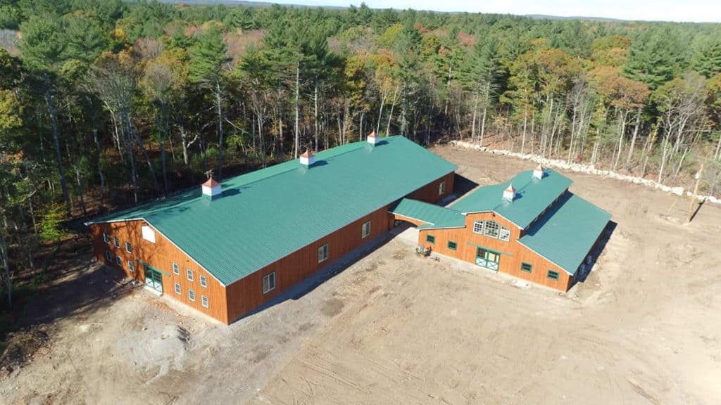 Aerial view of a large wooden barn with green metal roofing set in a forested area.