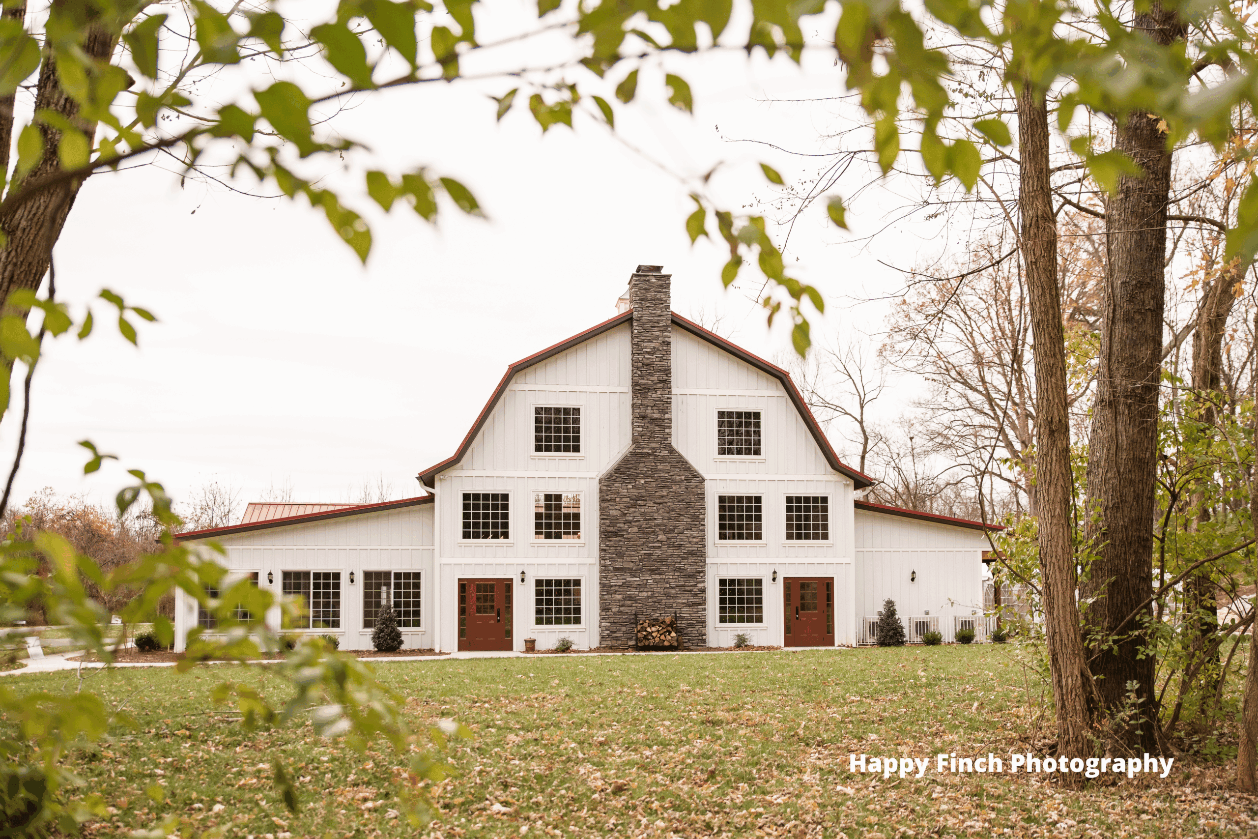 Barn building with rustic stone chimney, large windows, and white wooden siding, surrounded by trees.