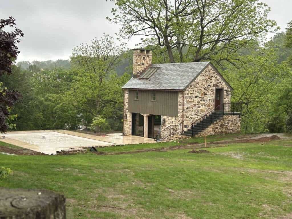 Detailed photo of a partially finished stone house with wooden accents, staircase, and surrounding greenery.