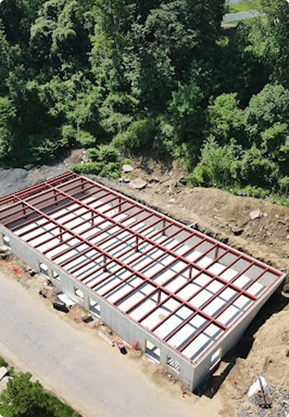Aerial view of a construction site showing a steel-framed commercial building in progress, with trees and natural landscape nearby.