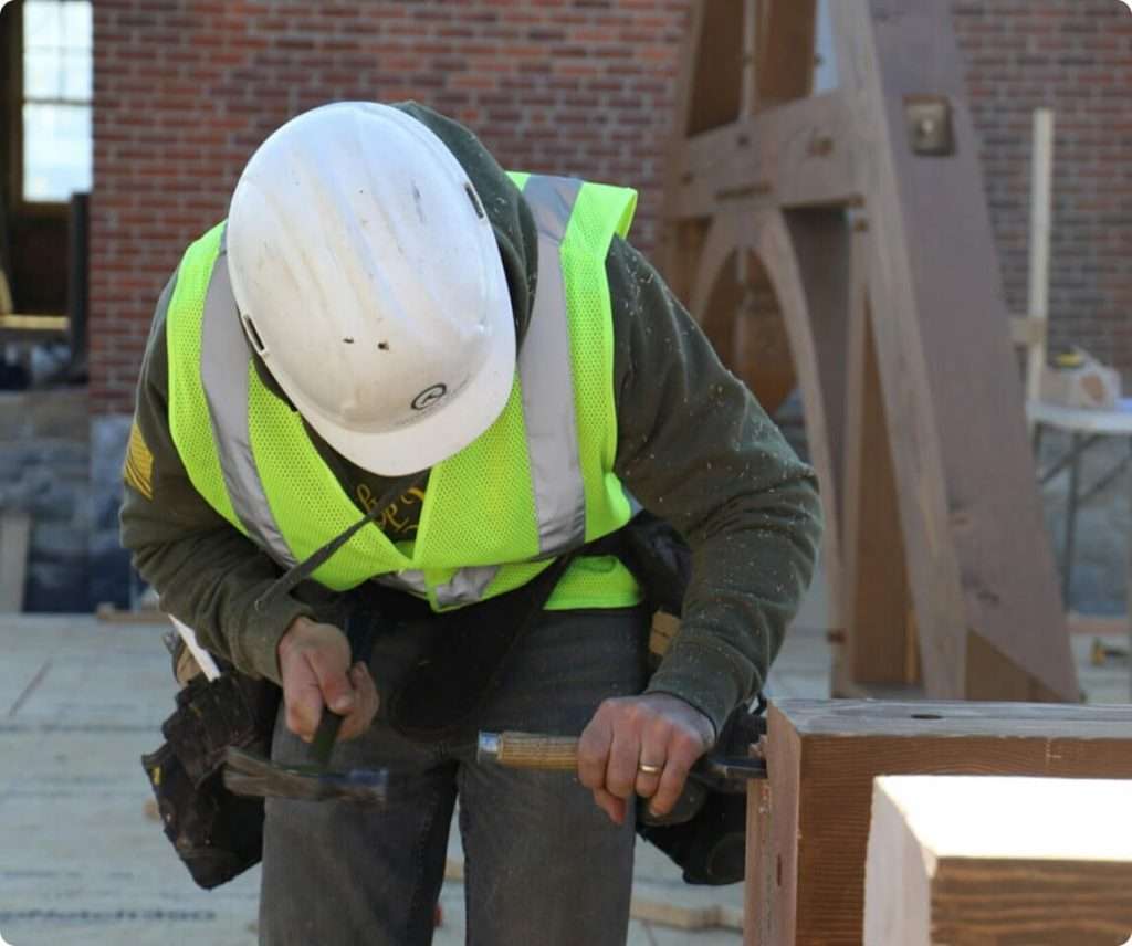 High-visibility safety vest and helmet on construction worker performing woodworking tasks at building site.