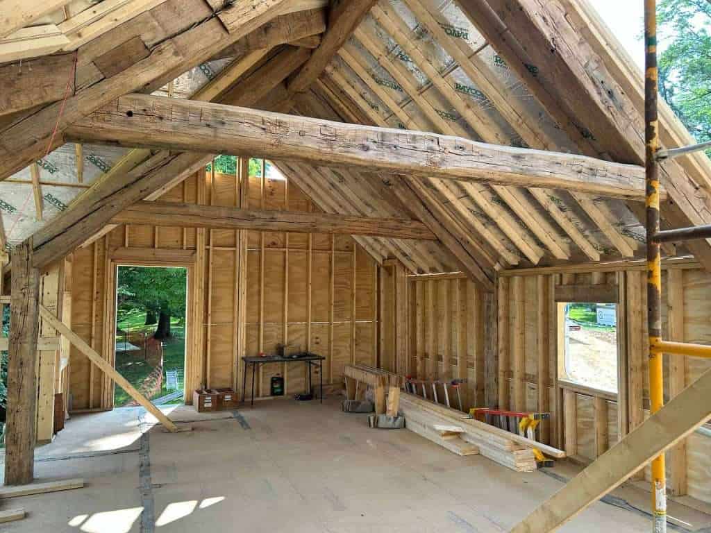Interior view of a wooden house under construction, featuring exposed timber framing, roof trusses, and construction tools.