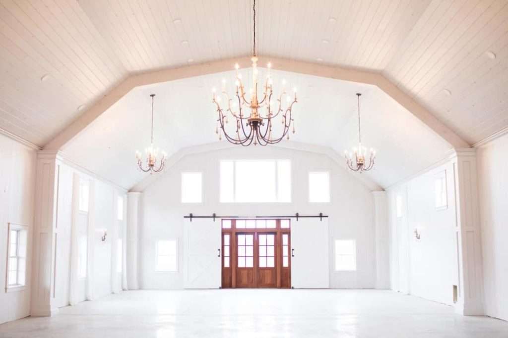 Spacious white barn interior with rustic chandeliers and large wooden entrance door.