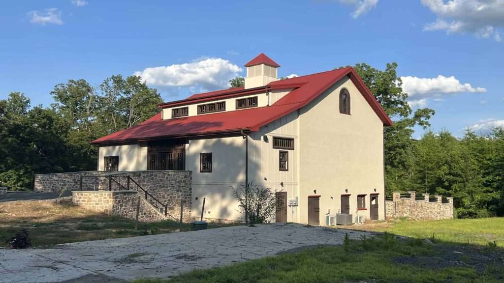 Rustic barn with stone foundation and red roof, nestled by trees under a blue sky.