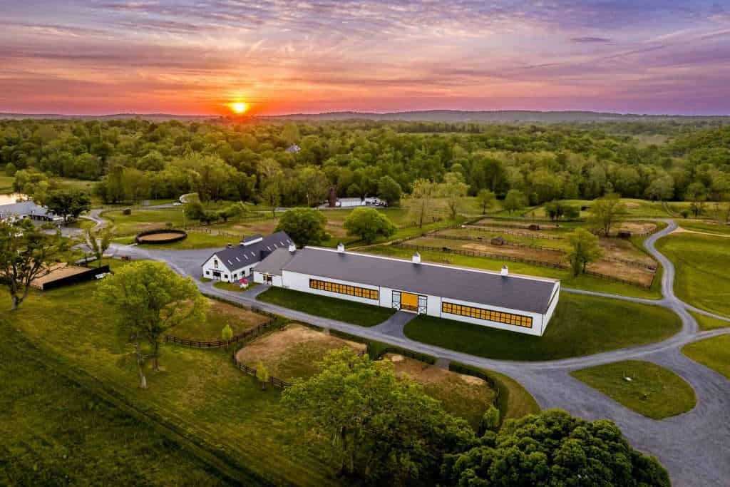 Aerial view of Quarry View Building Group's scenic property showcasing modern barn buildings, riding arenas, and lush green landscapes during sunset.
