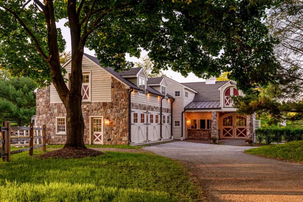 Full exterior view of a custom equestrian barn in West Chester, PA, at sunset, featuring matched stone masonry, cream siding, and red-trimmed carriage doors.